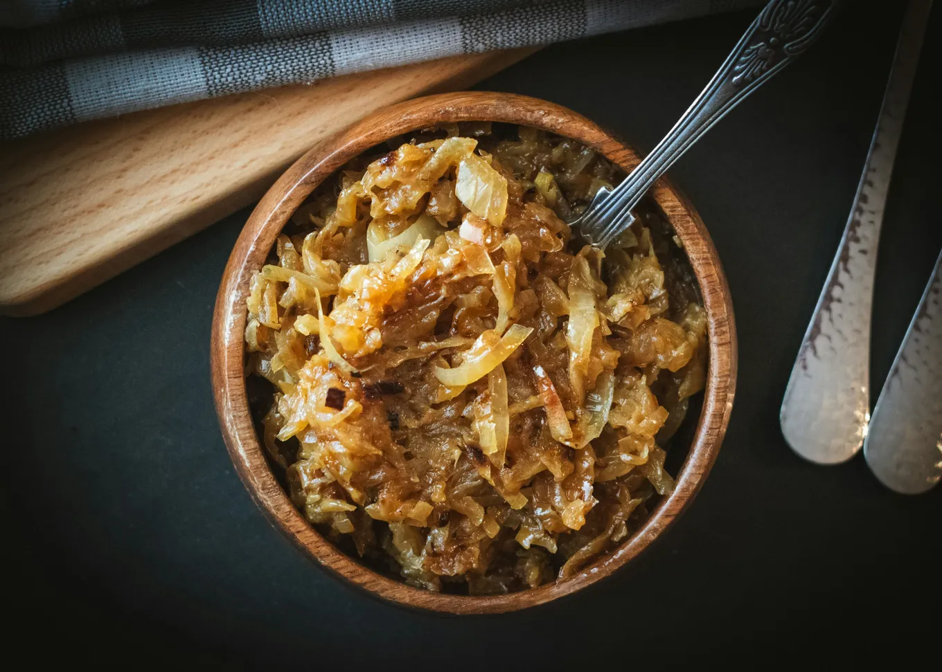 A wooden bowl of deep golden caramelized onions with a fork resting in it, on a dark surface beside a cutting board