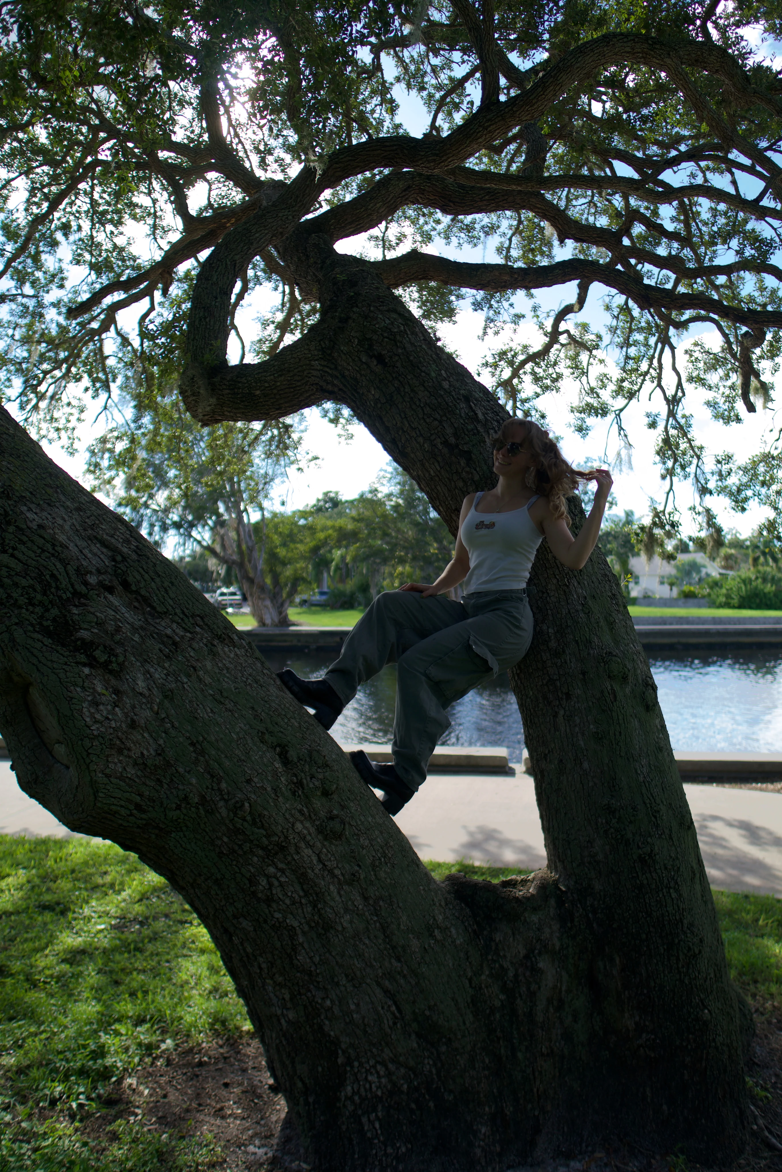 A woman leaning against the trunk of a leaning live oak beside a waterway, seen from a distance