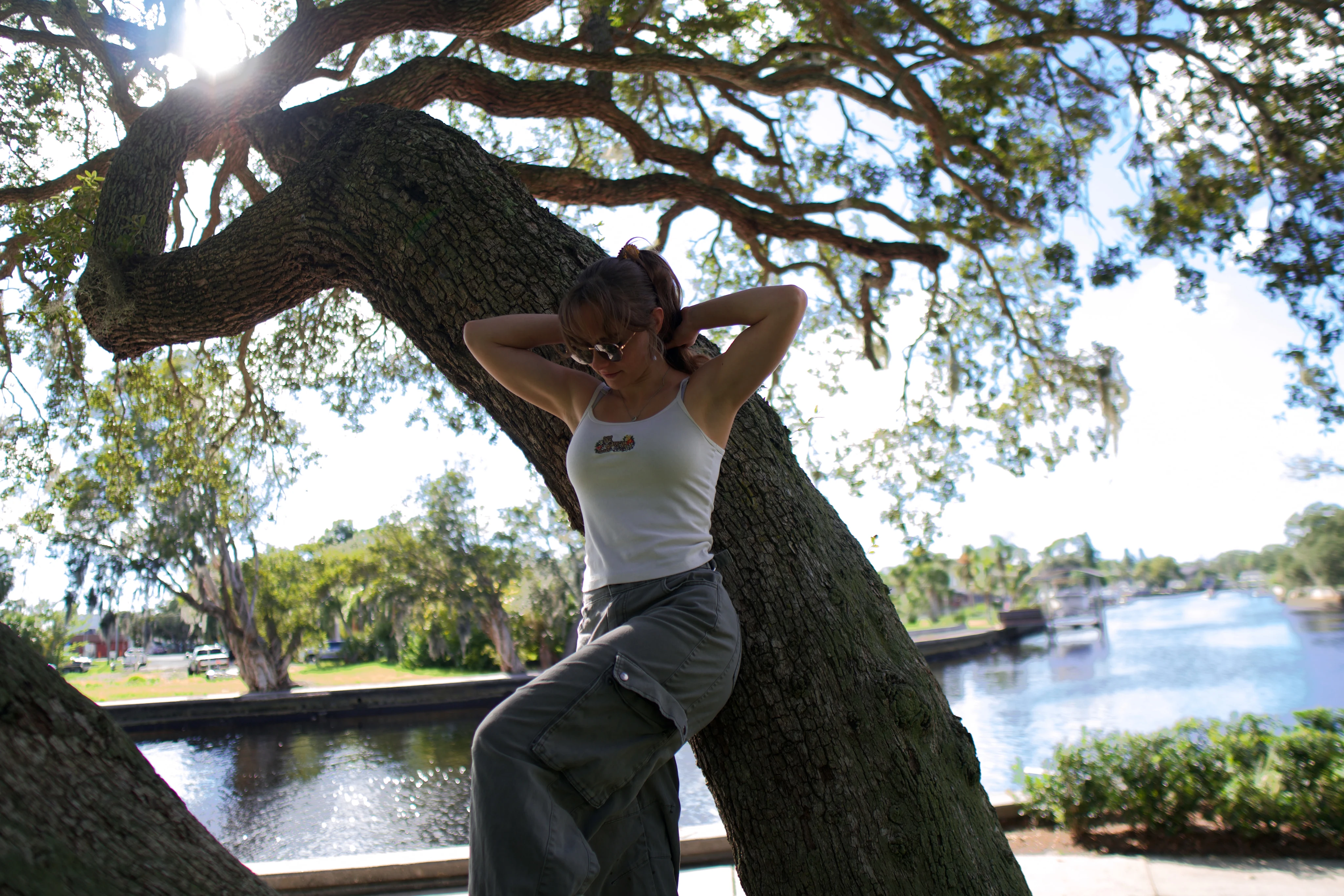 A woman sitting on a low oak branch with her hands behind her head, sun through the canopy