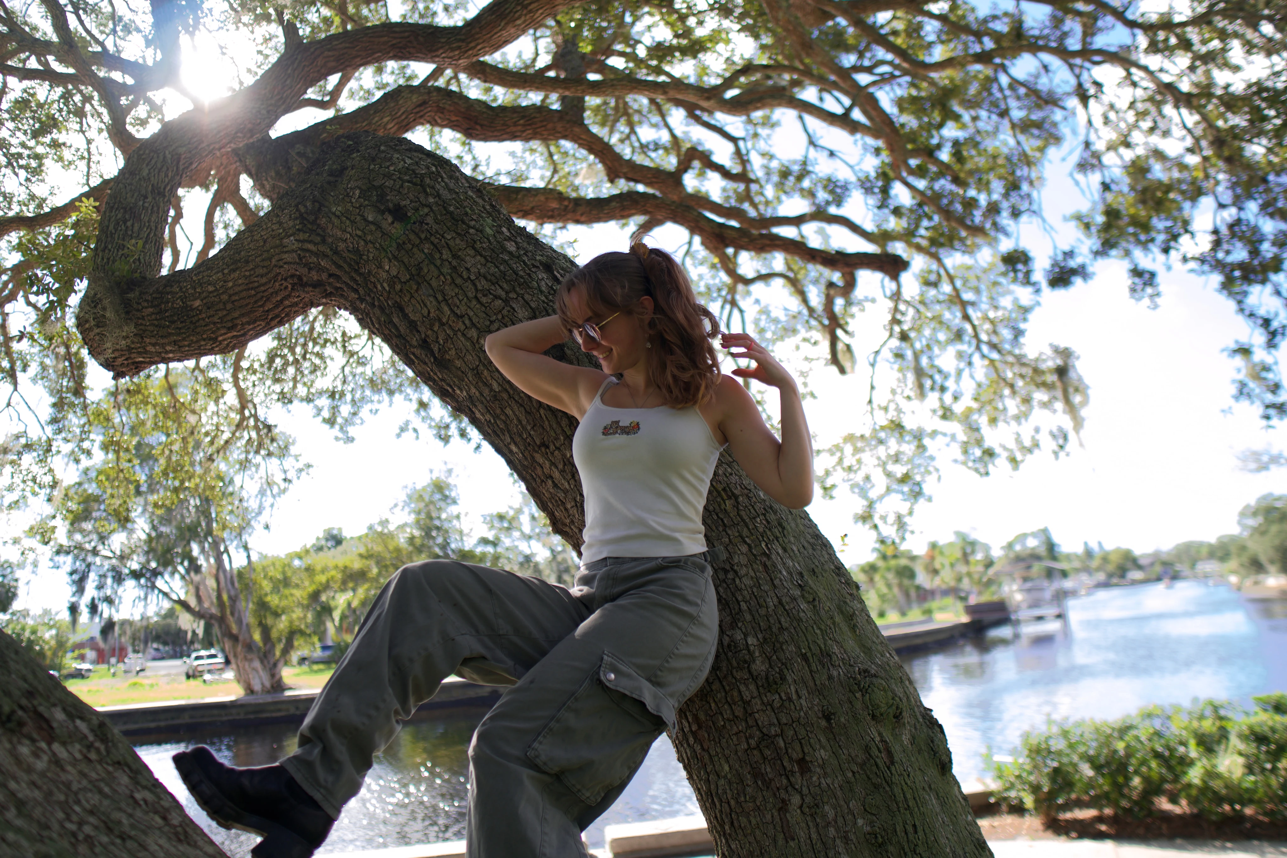 A woman seated on a leaning oak trunk with one hand in her hair, marina in the background