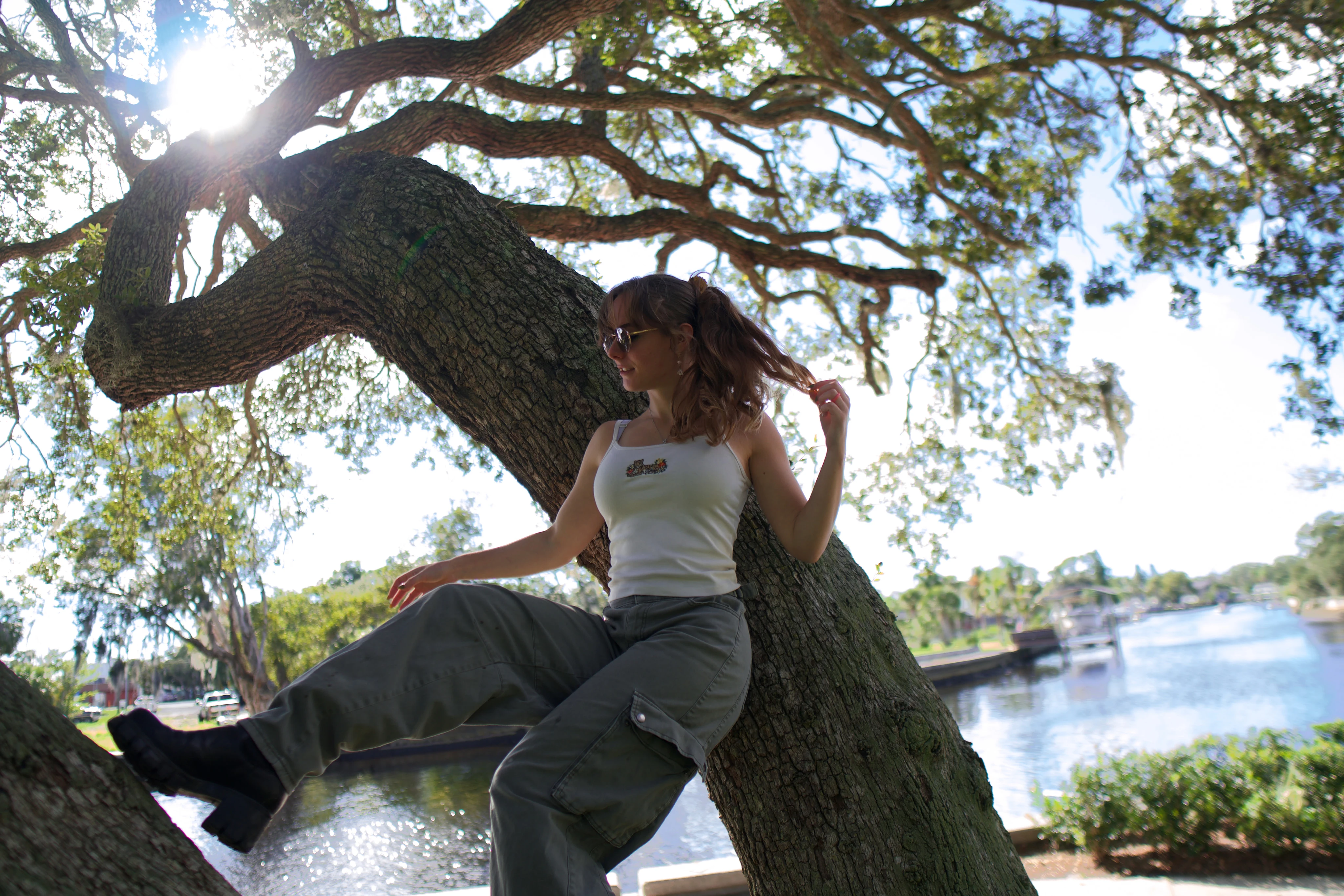 A woman seated on an oak trunk reaching out with one arm, sun flaring through the leaves
