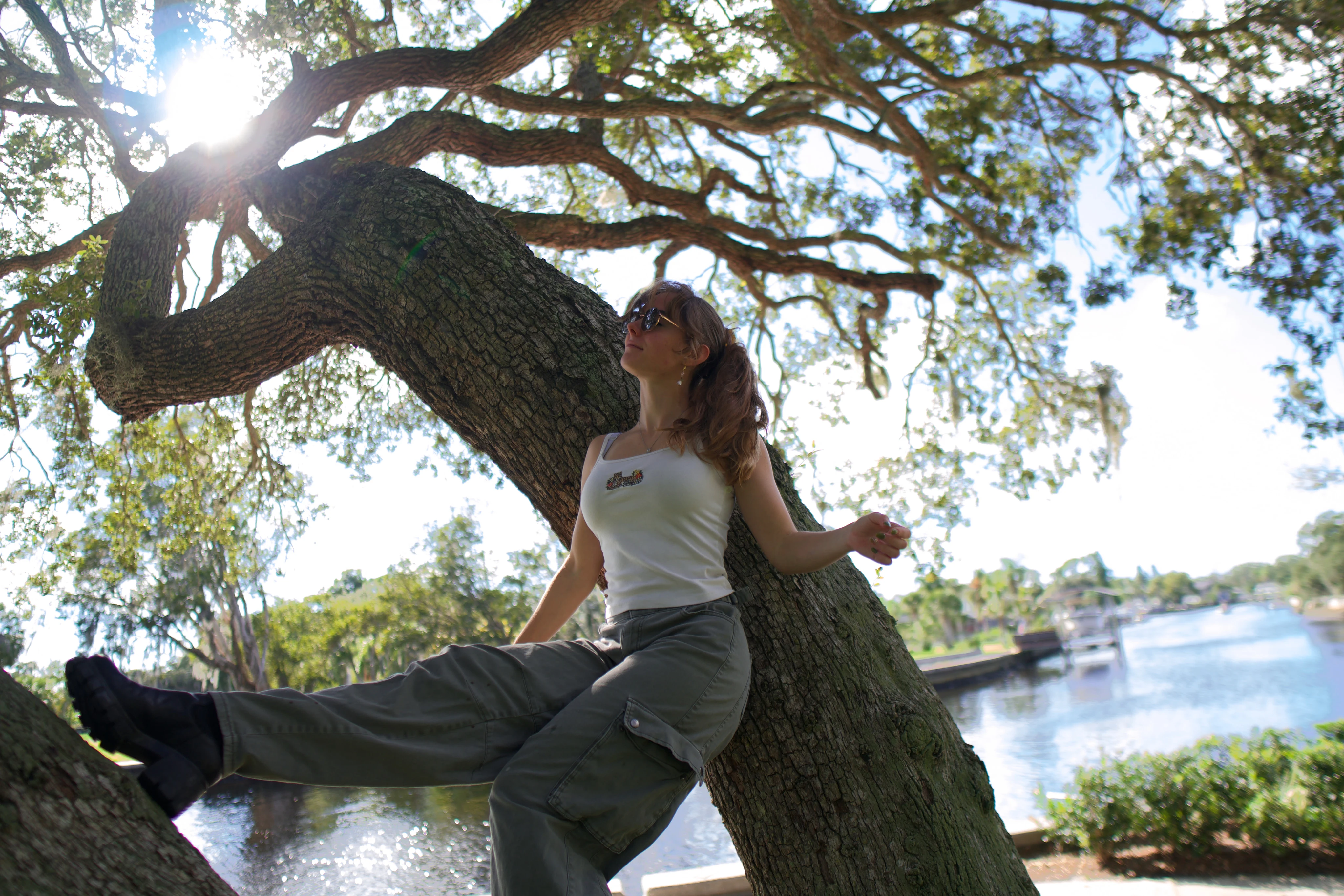 A woman seated on a leaning oak trunk, one arm outstretched, sunlit water behind
