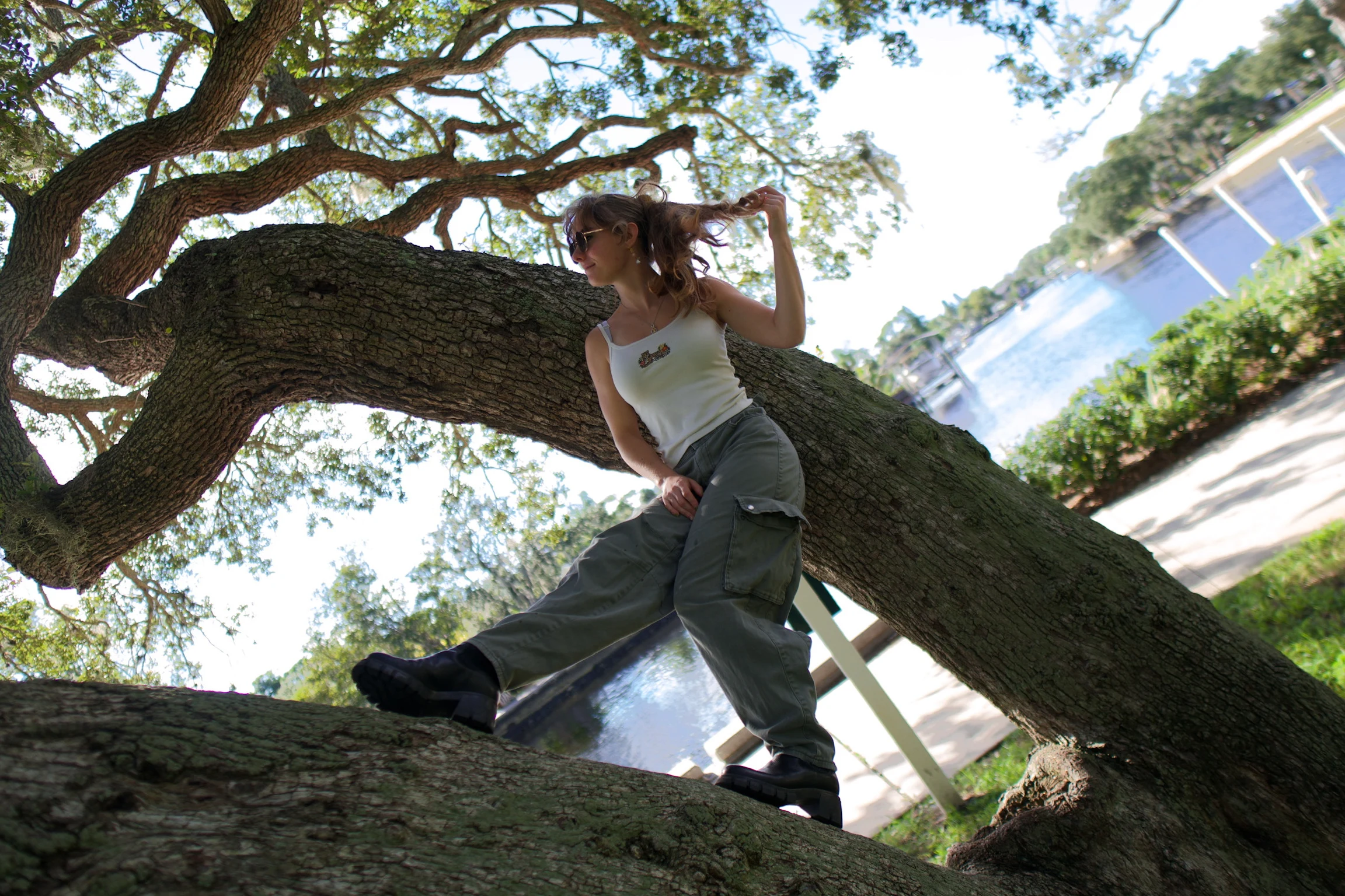 A woman standing on an oak trunk holding a branch overhead, blue sky through the leaves