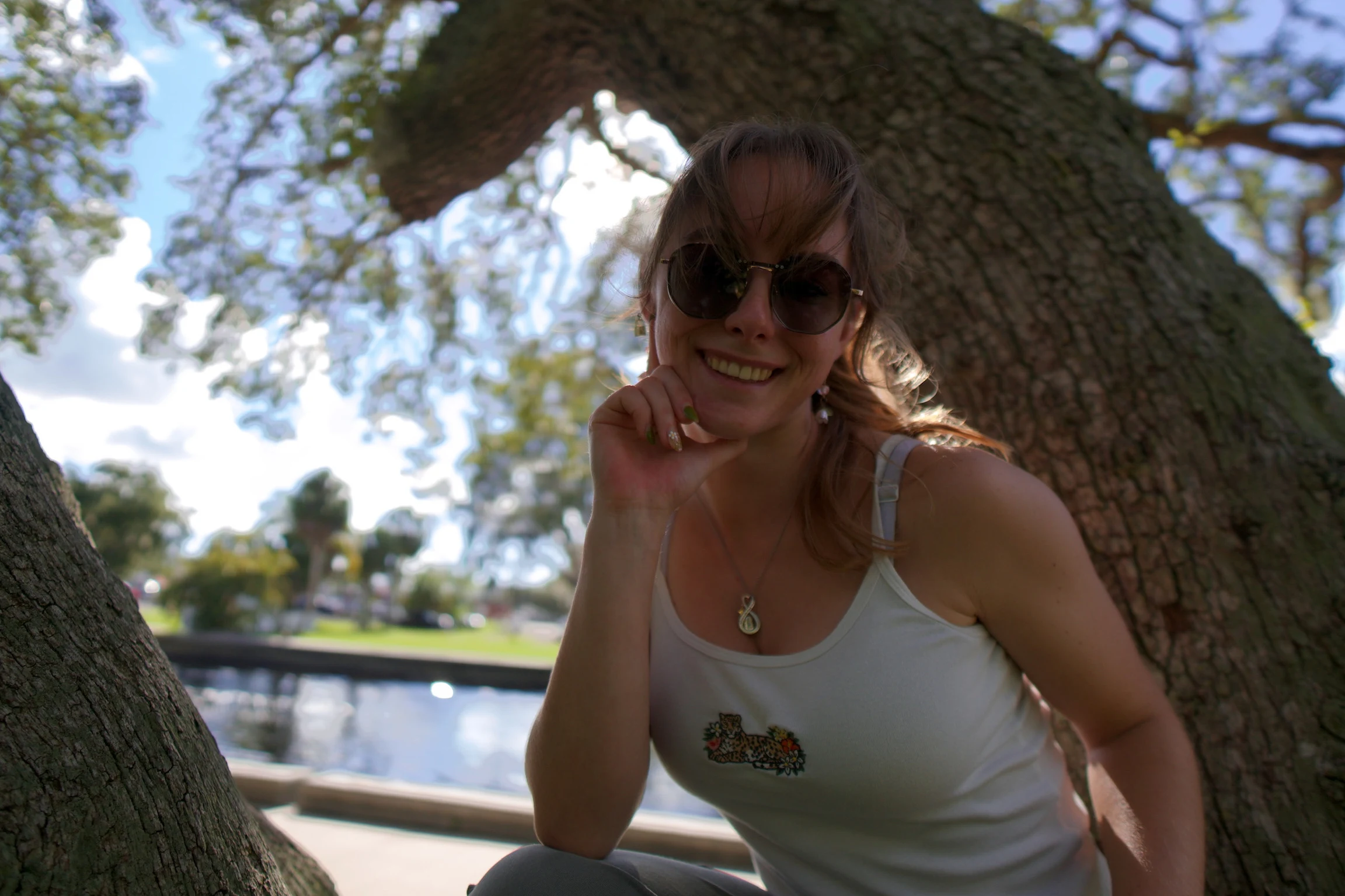 Close portrait of a woman in round sunglasses smiling under an oak, clouds behind