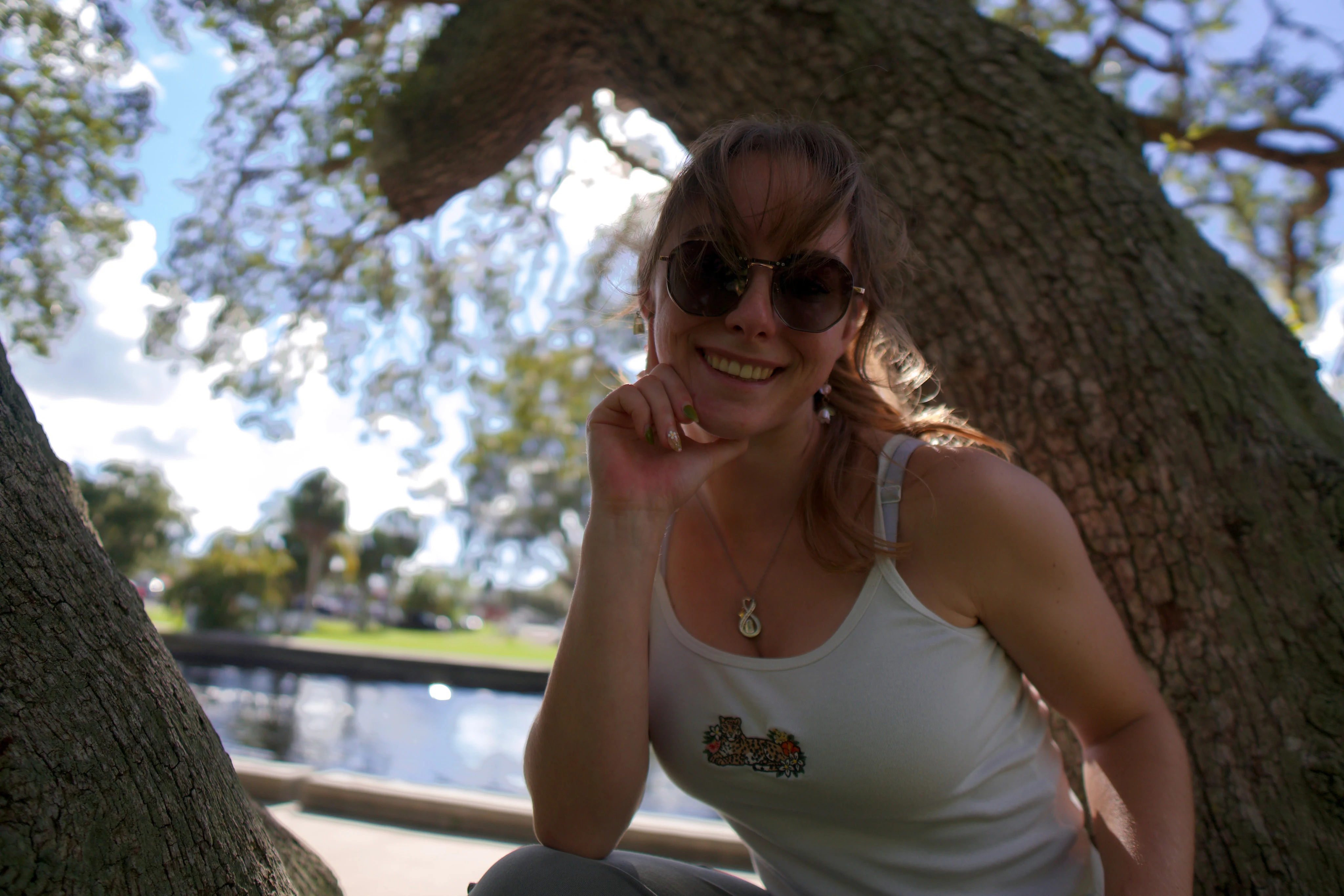 Close portrait of a woman in round sunglasses smiling under an oak, clouds behind