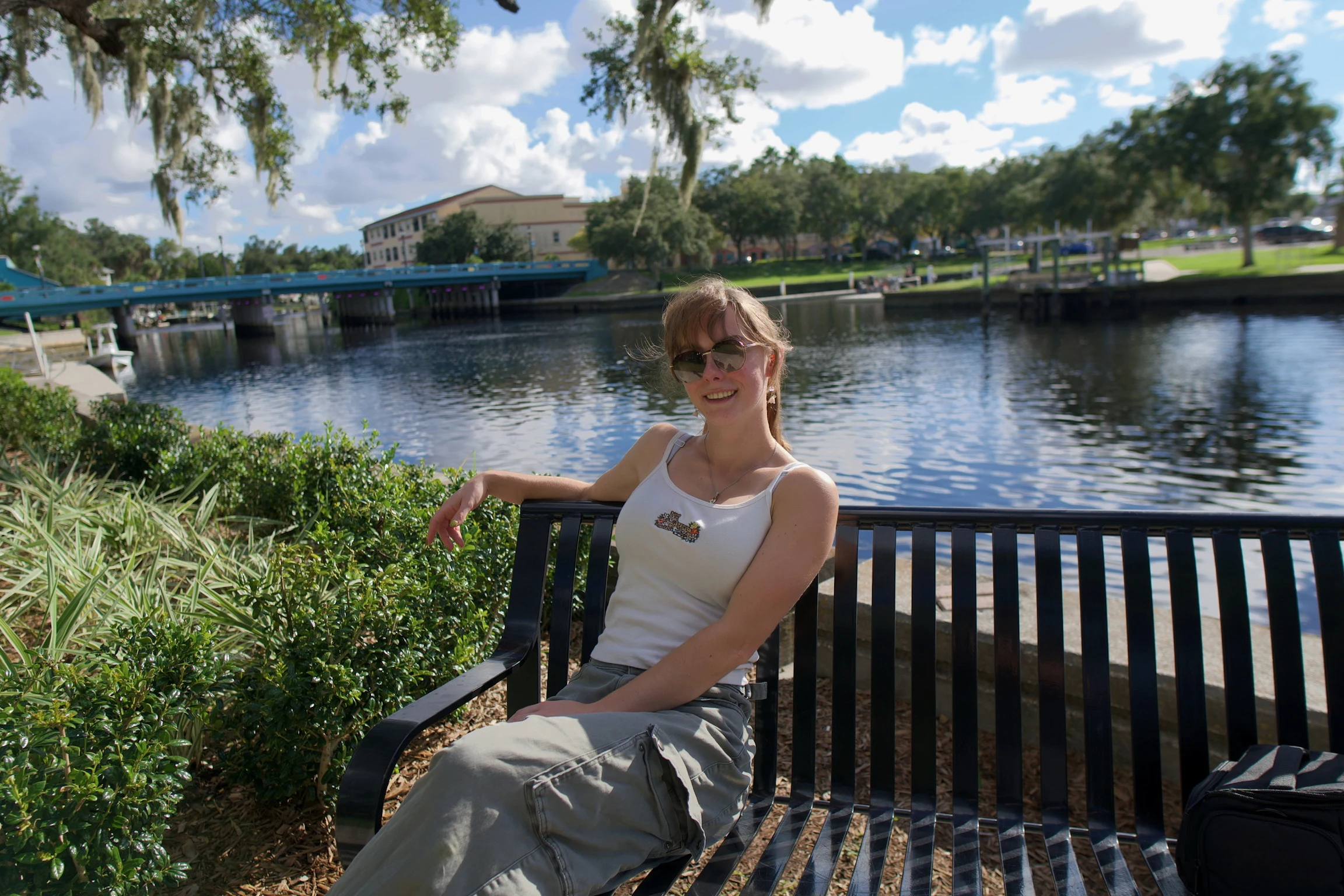 A woman sitting on a black metal bench by the water, arm along the backrest, clouds overhead