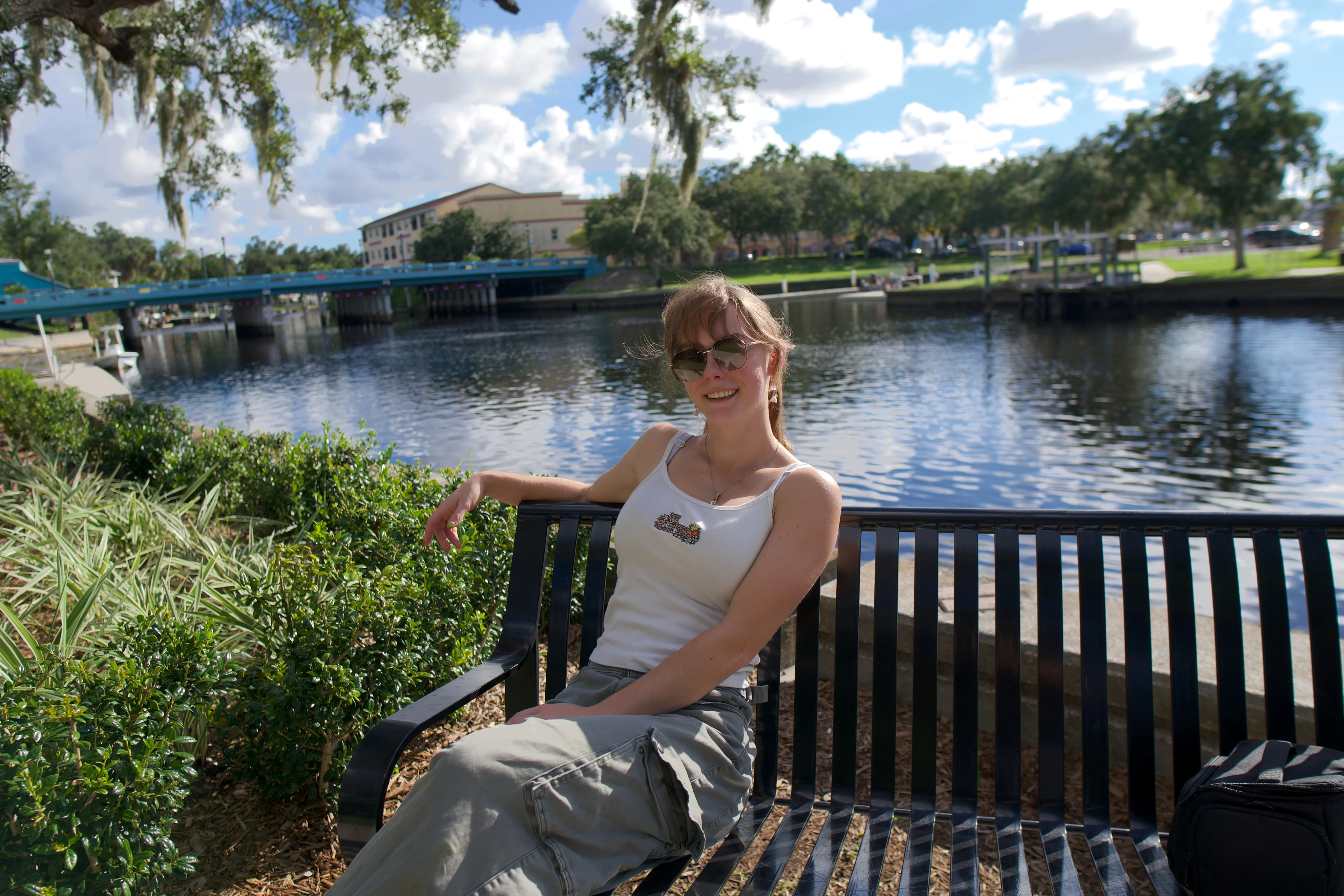 A woman sitting on a black metal bench by the water, arm along the backrest, clouds overhead
