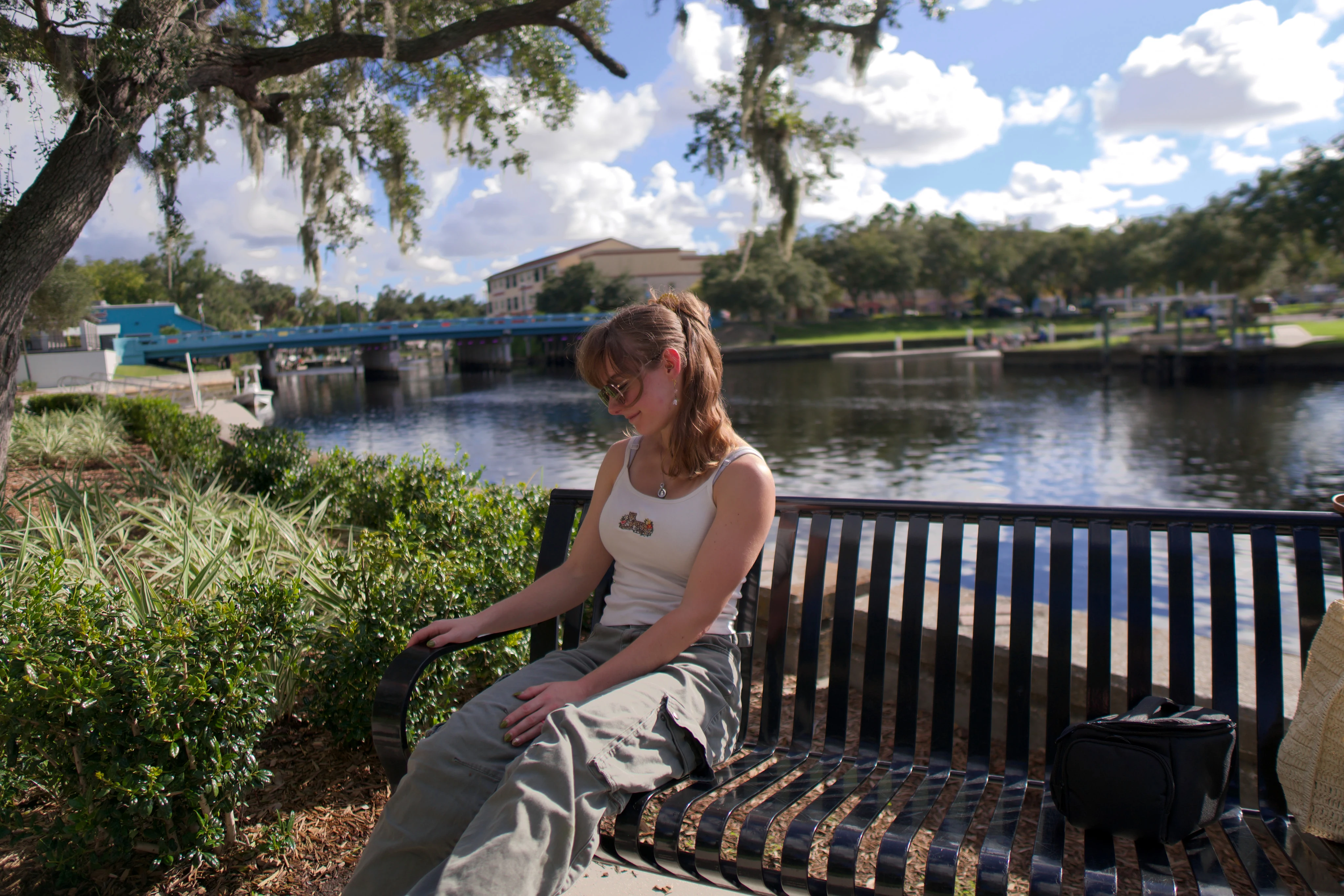 A woman on a bench by the water, looking down, blue sky and clouds