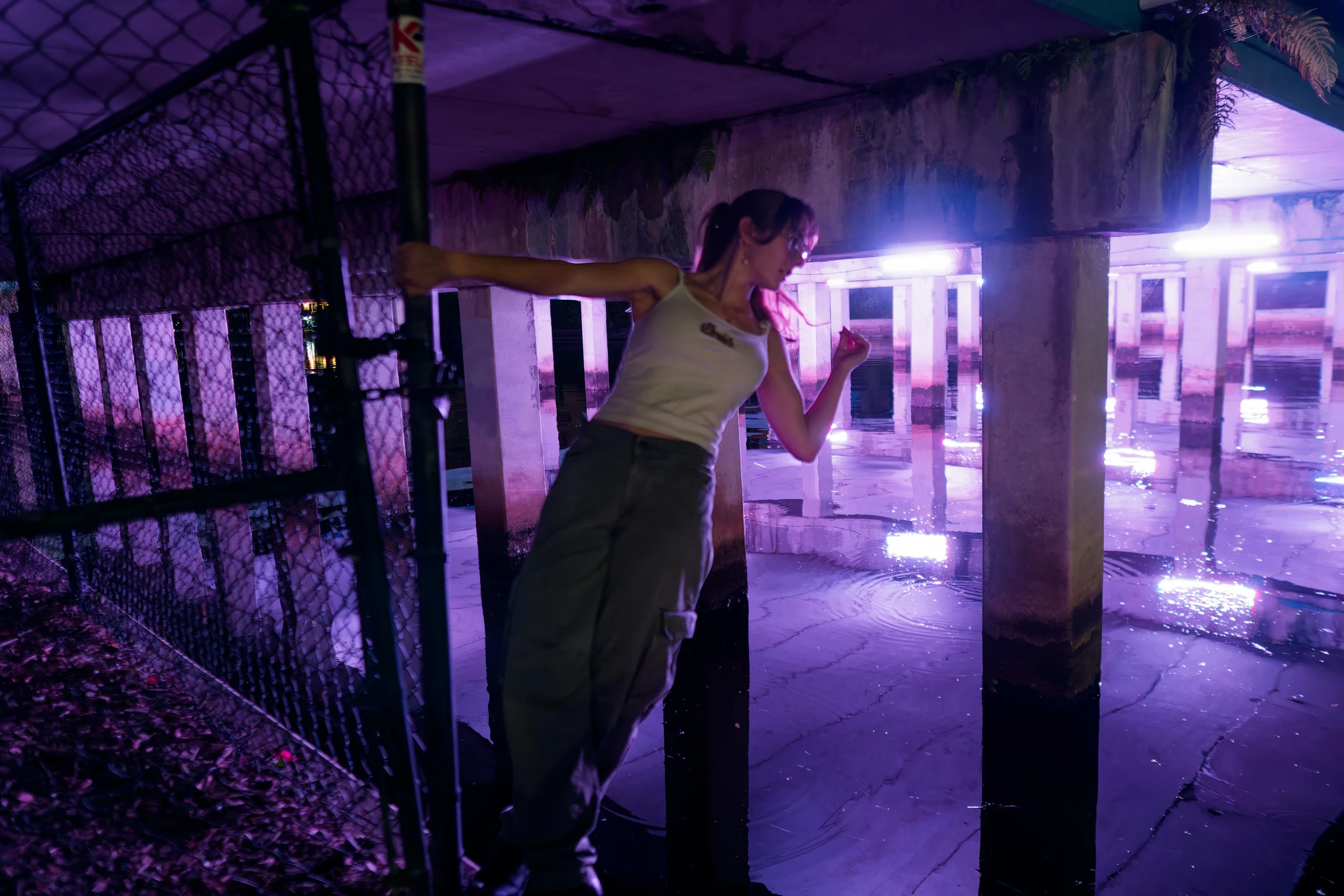 A woman leaning on a fence at night, looking away, purple-lit pier behind