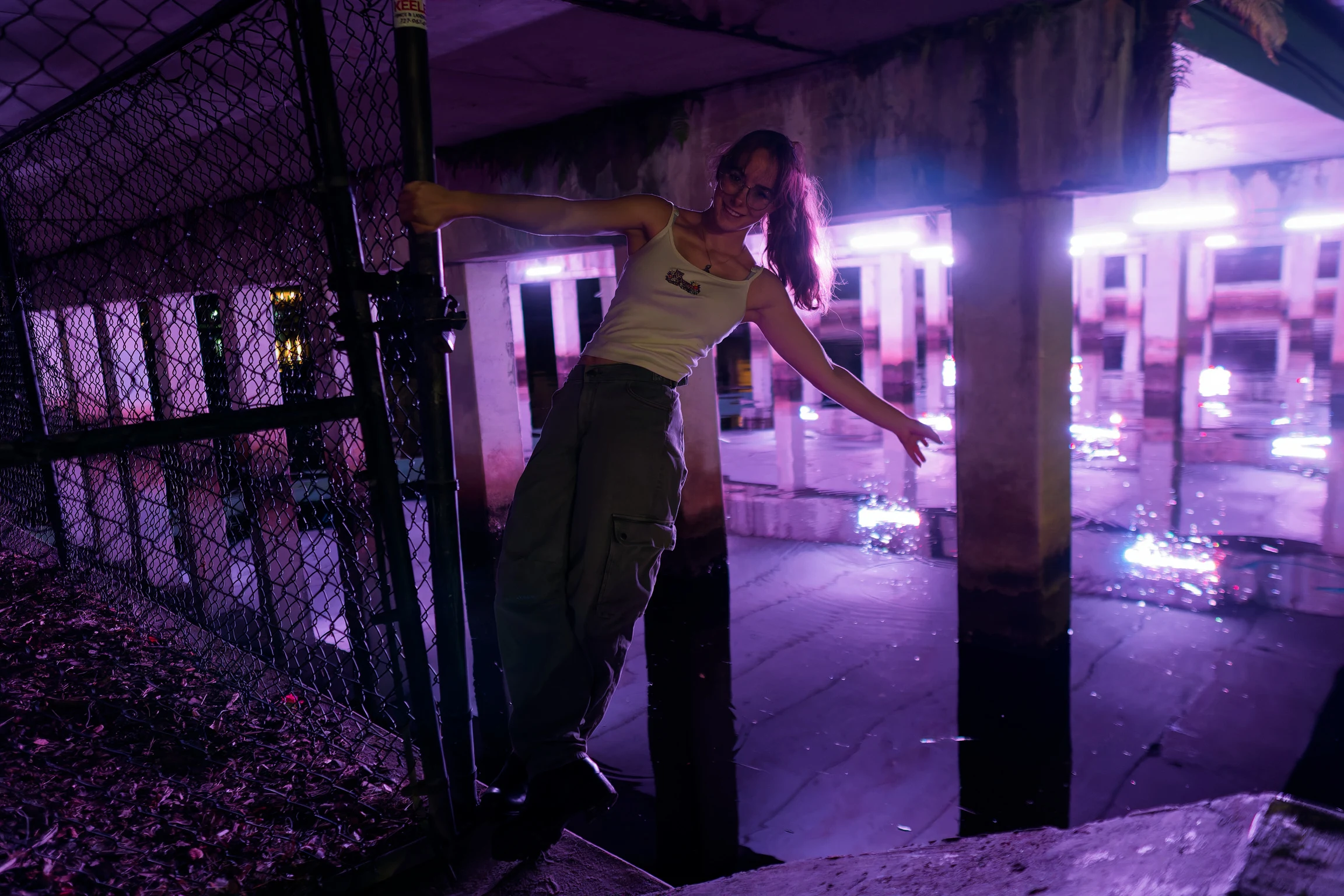 A woman at night with one arm outstretched along a fence under a purple-lit pier