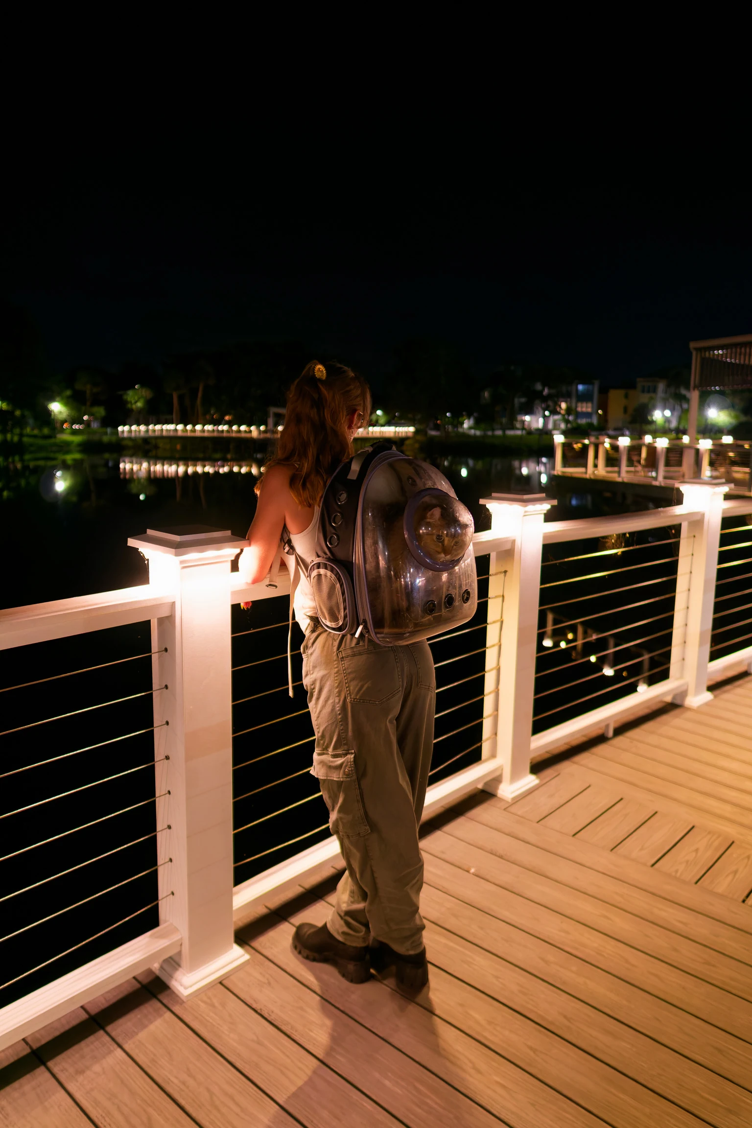 A woman with a backpack at a lit railing on a boardwalk at night, water beyond